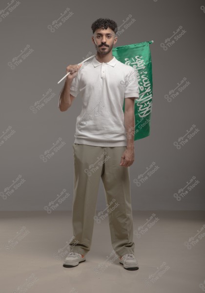 An image of a man standing and holding a green flag with white symbols, which is the flag of Saudi Arabia. He is wearing a white polo shirt and beige trousers with light-colored sneakers. The background is a neutral gray color.