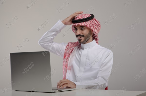 A photo of a man wearing traditional Saudi attire, a white thobe with a red checkered shemagh and agal, sitting in front of a laptop. He is smiling and placing his hand on his head, appearing surprised or pleased with what he sees on the screen.