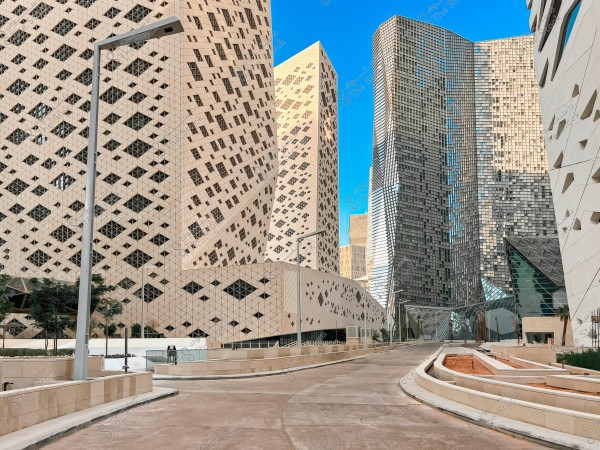 The image shows an urban scene with architecturally designed high-rise buildings in a modern area. The buildings feature unique architectural designs with latticework and glass facades. The foreground street is wide and free of vehicles, surrounded by modern pavements and lighting, reflecting a sophisticated urban atmosphere.