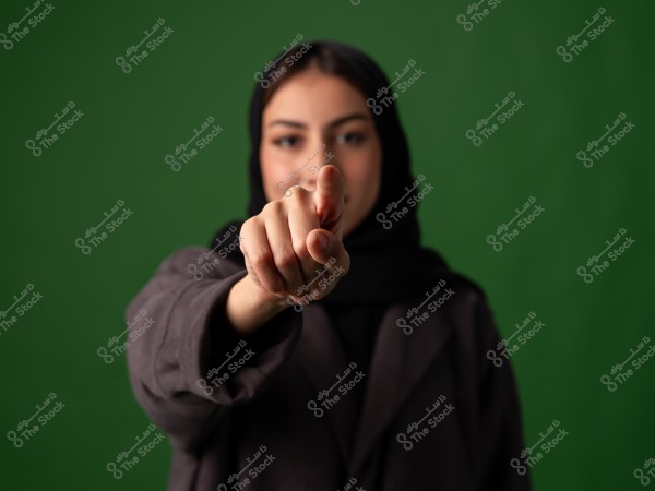 A portrait of a woman wearing a black hijab and a dark coat, pointing her finger towards the camera. The background is green.