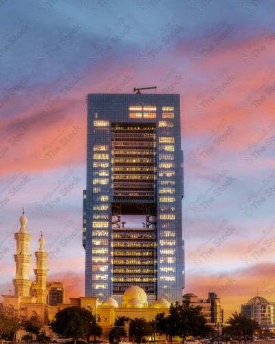 A picture of a building in the city of Riyadh, in front of it, a group of residential houses, showing the cloudy sky orange in color at sunset.