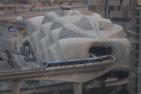A large white office building with curved, shell-like architectural design. A blue elevated train is passing in front without stopping and is visible at the forefront of the structure.