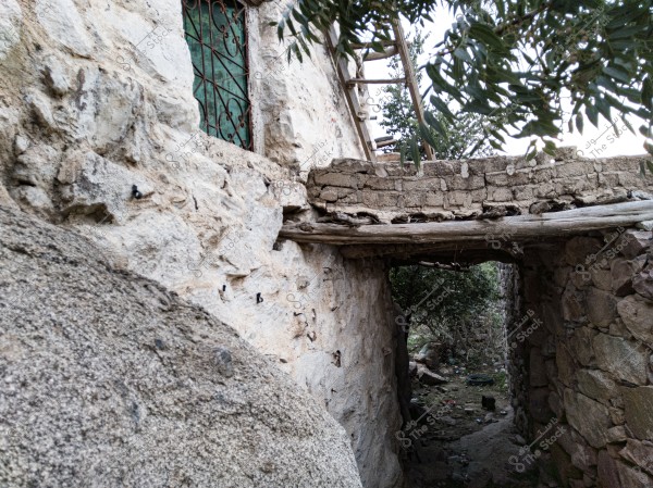 A photo of a narrow passage in a traditional stone building. Part of an old stone wall is visible, featuring a window with an iron frame covered by green glass. There is also an overhang made of bricks and wood, with a wooden ladder leaning against the wall. In the background, some trees and greenery can be seen.