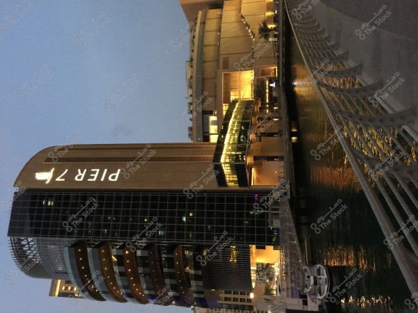 A tall building with an illuminated sign reading \"PIER 7\" is shown next to a waterfront marina. The building is lit with warm lights, and the light reflects on the water in the marina. A yacht is moored beside the building, and the sky appears dark blue, suggesting the photo was taken around sunset or night.