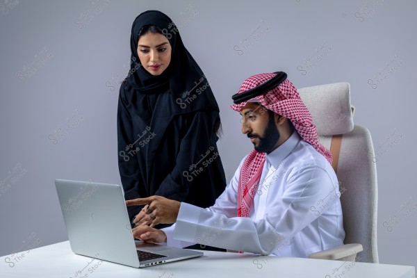A photo of a man and a woman in an office setting. The man is seated on a chair, wearing a white Saudi thobe and a red keffiyeh, working on a laptop. The woman stands next to him wearing a black abaya and hijab, appearing to point at something on the screen.
