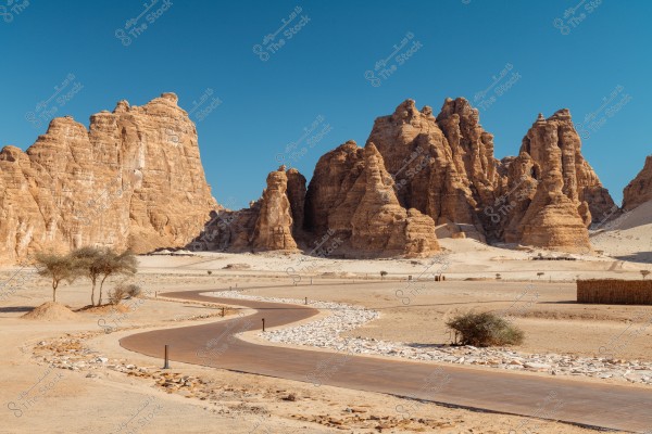 A landscape of a vast desert under a clear blue sky featuring a group of large, sand-colored rock formations. A paved, winding road passes through the foreground, with small desert trees and shrubs on either side. The rock formations in the background give the scene a majestic appearance.