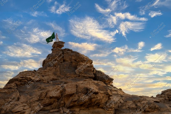 The image shows a person standing on the summit of a rocky formation in a vast natural landscape. The person is wearing traditional white clothing and holding a green flag. The sky is blue with scattered white clouds, giving a sense of openness and freedom. The scene is likely in Saudi Arabia, considering the design of the flag.