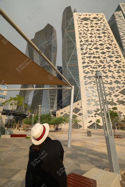 A photo showcasing modern skyscrapers with unique geometric designs, while a woman in a white hat with a red band sits on a wooden bench in the foreground. The scene includes scattered trees around the buildings, suggesting the image is taken in a contemporary urban area.