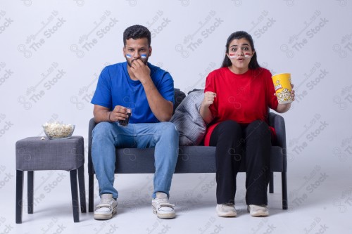 Portrait of a Saudi young man and woman watching a match and cheering their favorite teams cheerfully while having a snack on a blue background, the World Cup.