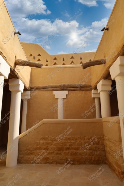 Image of a section of a traditional clay building featuring white columns and a ceiling adorned with timber beams. The sky is blue with clouds in the background, and the building reflects traditional architecture in Saudi Arabia.