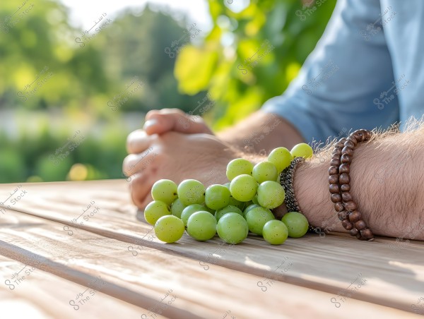 An image showing the hand of a person sitting at a wooden table in a green outdoor setting. The person is wearing a bracelet with wooden beads and a light blue fabric. A bunch of green grapes is on the table next to the hand.