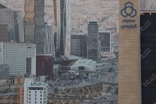 Image of Riyadh, the capital city of Saudi Arabia, featuring a collection of modern skyscrapers. In the center, there is a uniquely designed architectural building. There are also intricate roads with overpasses. On the right side, the logo of Al Rajhi Bank appears on a tall beige building.