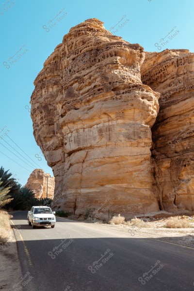 A white car drives on a paved road next to a massive rock formation in a desert area. The sky is clear and blue, and palm trees are visible in the background. Power lines can be seen at a distance from the road.