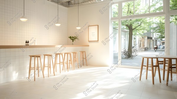 A modern, bright café with white tiled walls and long wooden tables surrounded by tall wooden stools. There is a glass vase with green plants on the bar table. Light streams in through large glass windows overlooking the outside street, which features trees and parked bicycles. The ambiance is calm and minimalist with warm natural lighting.