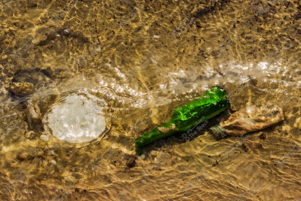 Image showing a green bottle and a plastic cap partially submerged in clear, sandy water. Light reflections are visible on the water's surface, with subtle details of the sandy ground beneath.