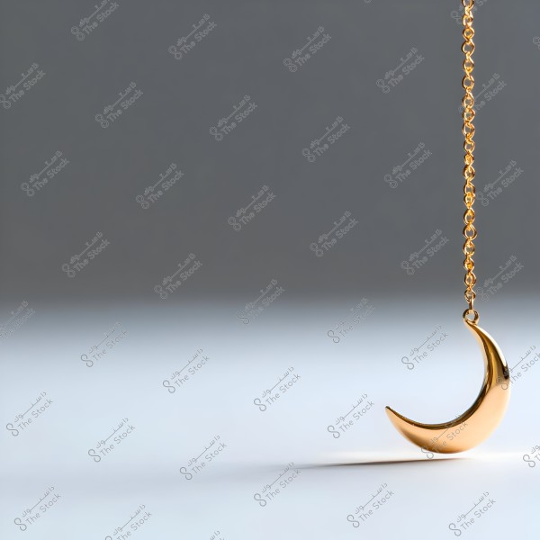 A gold chain hanging in the air with a shiny gold crescent pendant. The background is blurred with a gradient from gray to white, highlighting the luster of the metal.
