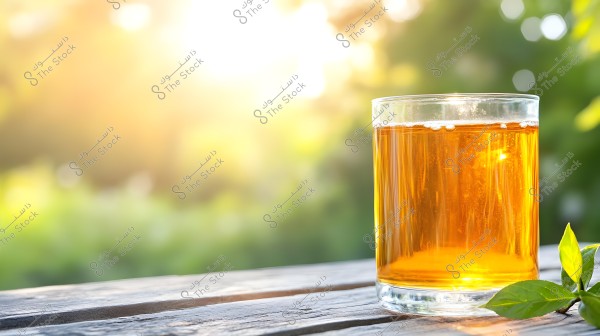An image of a transparent glass filled with golden-colored tea placed on a wooden outdoor table. The background is blurred, featuring sunlight filtering through trees, giving a warm and natural feeling. Some green leaves are seen next to the glass.