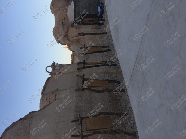 The image shows part of an archaeological site with a wall made of natural rock. The wall is adorned with wooden plaques mounted on clay structures. A stone-paved path runs alongside the wall. The sky is clear blue in the background.