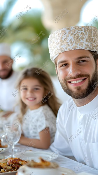 A man wearing an Omani dishdasha and kumma sits smiling next to a young girl in a white dress. There is a spread of food on the table in the foreground, with another person partially visible in the background. The background includes green plants.