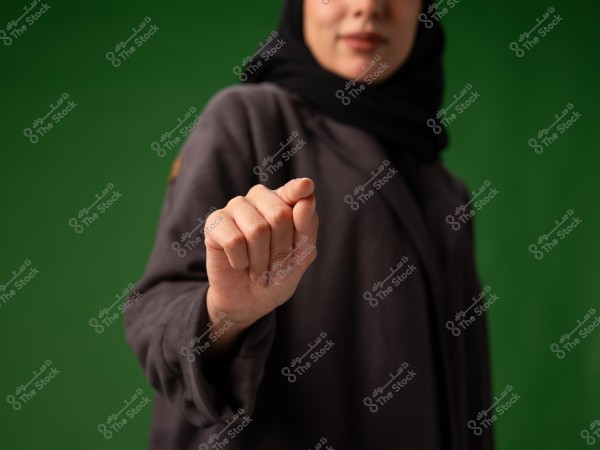 تظهر الصورة سيدة ترتدي حجابًا, تُركز على يدها وهي ممدودة على خلفية خضراء.