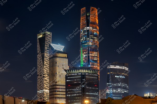 A view of the Riyadh city skyline at night, featuring a cluster of modern skyscrapers illuminated with colorful lights. The towers display unique and diverse architectural designs, adding an urban and innovative atmosphere to the nighttime scene.
