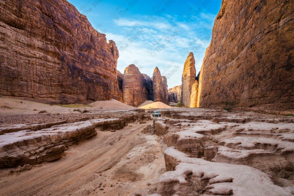 لقطة لسيارة وسط تجاويف صخرية في محافظة العلا محاطة بسلسة من الجبال الصخرية وتظهر السماء صافية نهارًا، الطبيعة بالسعودية، اماكن سياحية.