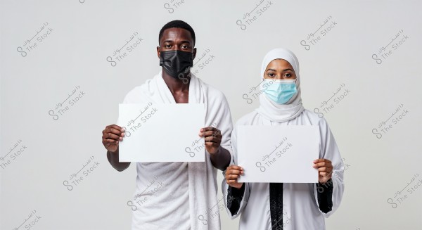 A photo of a man and a woman standing side by side, both wearing face masks. The man on the left is dressed in simple white clothing and is holding a blank board. The woman on the right is wearing a hijab and a white robe adorned with black decoration, also holding a blank board. The attire appears to be traditional as seen in the Saudi context.
