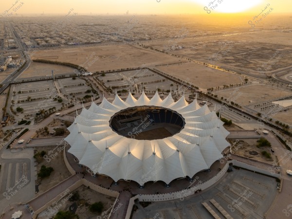 The image shows a large stadium with a circular design covered by a white, tent-like roof. In the background, there is a vast urban landscape characterized by desert and buildings, with the sunrise on the horizon providing a beautiful golden light to the scene.