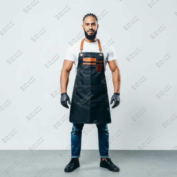 An image of a man standing upright on a gray floor with a white background. He is wearing a fitted white shirt, a black apron with brown leather details, and black gloves. He has blue jeans and black shoes. The man appears to be working in a craft or culinary profession.