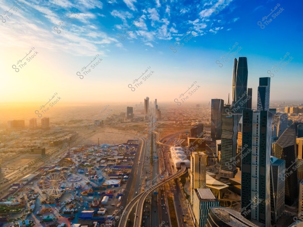 صورة جوية لمدينة الرياض في المملكة العربية السعودية. تظهر في الصورة مجموعة من ناطحات السحاب الحديثة والمباني الشاهقة التي تشكل أفق المدينة. في الجانب الأيسر من الصورة, تُظهر المتنزهات وأماكن التجمعات المجاورة للطريق الرئيس الذي يمر عبر المدينة. تظهر السماء صافية وأشعة الشمس تفيض من جهة اليسار, مما يضفي لمسة دافئة على المشهد.