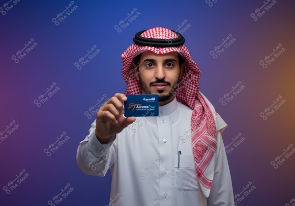 A portrait of a man wearing traditional attire with a red checkered headscarf, holding a blue card that says \"Alinma,\" against a gradient blue to brown background. He appears to be Middle Eastern, possibly from Saudi Arabia.