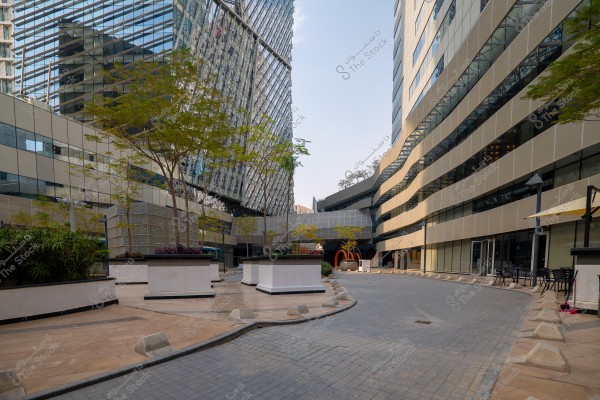 An image of a modern outdoor space between tall glass buildings. The scene shows a paved pathway leading towards a series of buildings with sleek architectural design and sharp angles. Trees and greenery adorn the area, while there are some outdoor seating and tables. The glass facades add a contemporary and sophisticated touch to the setting.