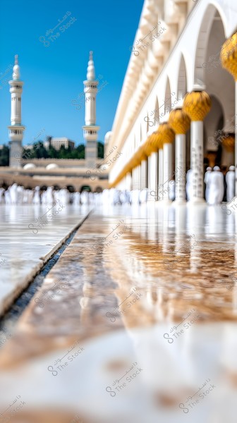 An image showing a part of the Masjid al-Haram in Mecca, Saudi Arabia. The picture features tall white minarets and the facade of the mosque with a line of arches and golden lanterns, with pilgrims in white ihram garments walking on a shiny marble surface reflecting the light.
