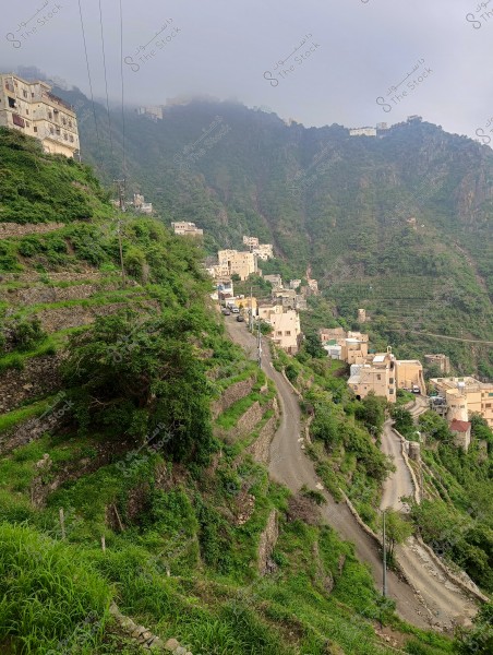 A lush mountainous terrain with winding roads and a cluster of residential buildings on the mountain slope. Clouds cover the mountain peaks, creating a misty atmosphere. Vegetation thrives on the agricultural terraces surrounding the area.