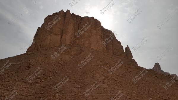 View of a large rocky mountain in a desert area under a slightly cloudy sky, with the sun shining from behind, highlighting the brown rock details. The ground is covered with gravel and dirt, with extensions of the mountain visible in the distance.