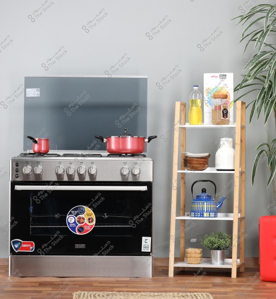 An image of a kitchen featuring a large silver stove with two red pots, one small and one large, on top. Next to the stove, there is a wooden shelf with four tiers holding various items like a bottle of oil, a box of cornflakes, a ceramic jug, a set of wooden bowls, and a small plant pot. The background has a grey wall and an attractive plant in the corner of the room.