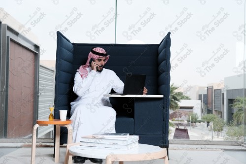 رجل سعودي يرتدي الزي السعودي التقليدي يقوم بإجراء مكالمة هاتفية بينما يدون ملاحظاته على إحدى صفحات المفكرة ويظهر الجهاز المحمول على سطح الطاولة أثناء النهار