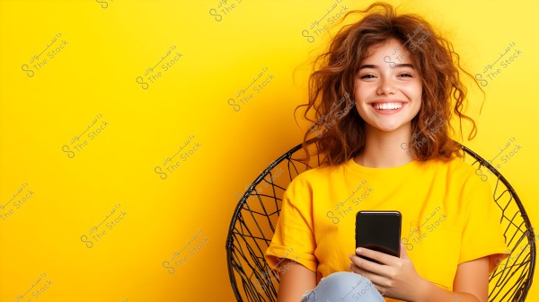 A portrait of a young woman sitting and smiling on a wire chair against a bright yellow background. She is wearing a yellow T-shirt and holding a smartphone in her hand. Her hair is brown and naturally curly.