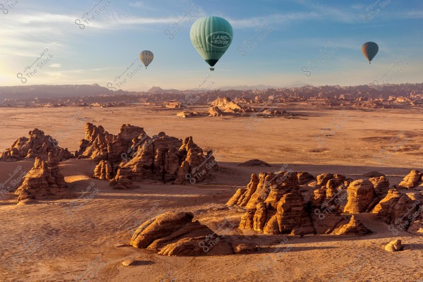A desert landscape showcasing unique rock formations with three hot air balloons flying above. The largest balloon bears the \"LAND ROVER\" logo. The sky is clear and light blue, and the rock formations are bathed in golden sunlight.