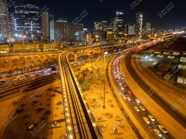 Night scene of a modern city showcasing tall skyscrapers illuminated with bright lights. A central elevated highway bridge crosses over heavy traffic, with the taillights of vehicles appearing as bright streaks. Palm trees are scattered on both sides of the road, with a landscaped area surrounding the highway.