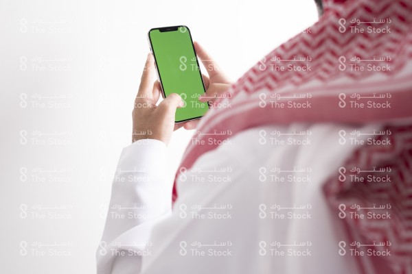 صورة ملتقطة بهاتف من الخلف لرجل سعودي يرتدي الزي السعودي التقليدي يستخدم الجوال وتظهر شاشة الجوال بوضوح