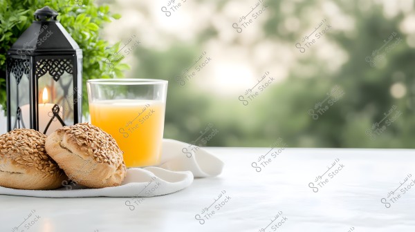 An image of a table featuring seed-covered bread rolls and a glass of fresh orange juice next to an illuminated traditional lantern on a white table. Green plants are in the background.