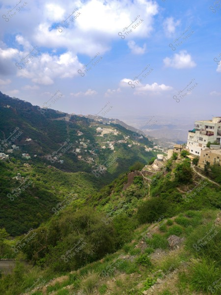 منظر طبيعي لجبل أخضر مغطى بالأشجار مع مباني بيضاء تقف على حافته. تمتد طريق متعرجة عبر المشهد, وتظهر في الخلفية جبال أخرى بعيدة تحت سماء زرقاء مزينة بغيوم قليلة.