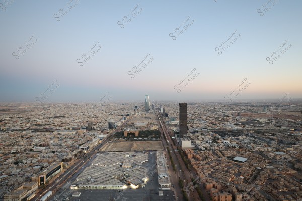 Aerial view of Riyadh, Saudi Arabia, featuring the Kingdom Tower at the center under a clear blue sky. Roads and streets extend in all directions, surrounded by bustling buildings and cityscape.