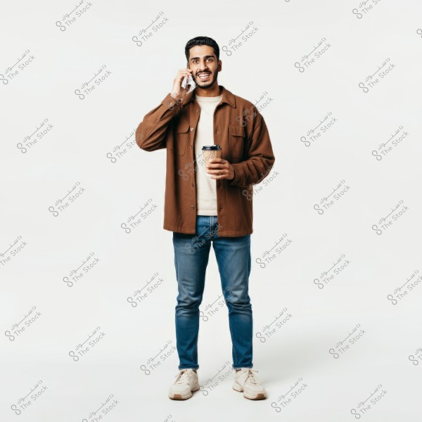 An image of a man standing and smiling while talking on a mobile phone, holding a paper coffee cup in his other hand. He is wearing a brown shirt over a light t-shirt, blue jeans, and beige sneakers. The background is completely white.