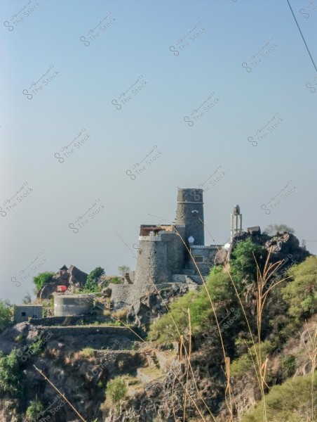 The image shows a traditional stone building on top of a hill surrounded by greenery and trees. The building features a circular tower and thick walls, resembling an old castle or fortress. In the background, the clear blue sky is visible.