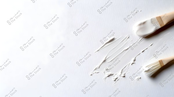 Two light-colored brushes placed on a surface painted in white. The paint is spread in smooth lines by the brushes, giving the image an artistic texture.