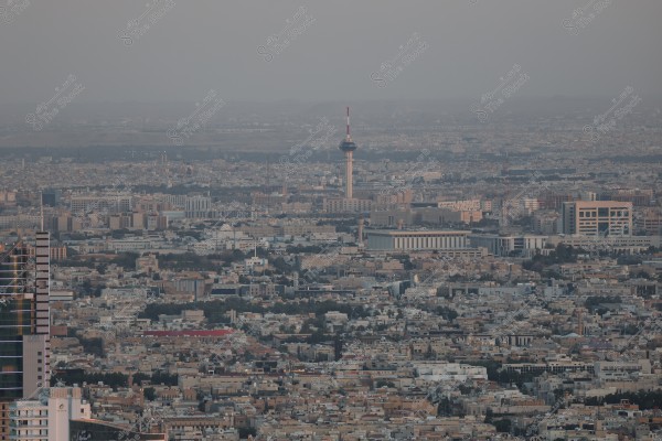 Aerial view showing a wide urban landscape of Riyadh city, with modern and diverse buildings spread throughout. The Riyadh TV Tower is prominently visible in the background, surrounded by numerous residential and commercial buildings, along with interconnected roads. The skyline is enhanced with the onset of sunset, casting a warm ambiance over the city.