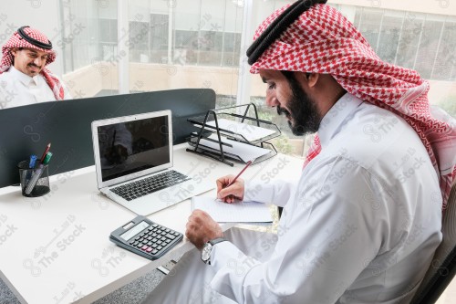 رجل سعودي يرتدي الزي السعودي التقليدي يعمل في مكتب بواجهة زجاجية ويستخدم قلم رصاص لتدوين الملاحظات