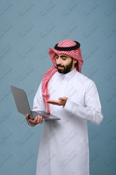 بورتريه شاب سعودي يرتدي الزي السعودي يأشر إلى جهاز المحمول وتظهر على ملامحة السعادة ، يقف على خلفية سماوية